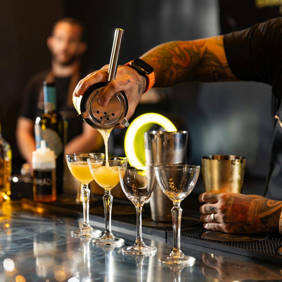 Bartender pouring a cocktail into glasses at a bar, with blurred bartender in background and various bar tools and bottles on the counter.