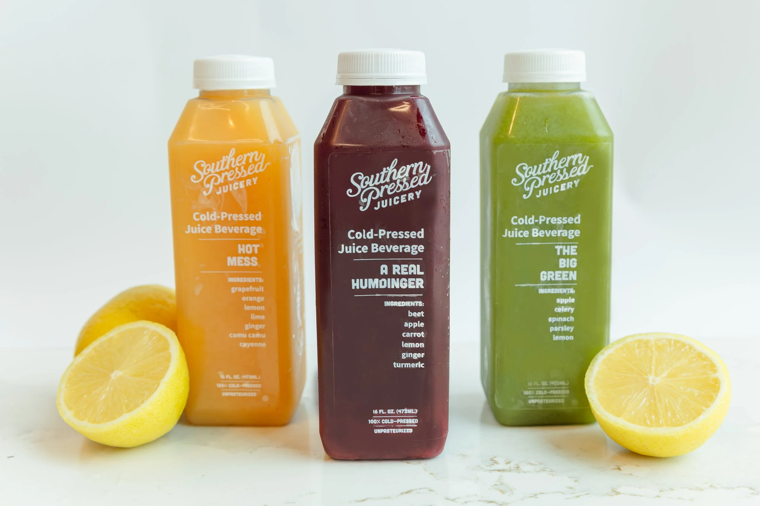 Three bottles of cold-pressed juice from Southern Pressed Juicery in front of lemon halves on a white surface. The juice flavors are Hot Mess, A Real Humdinger, and The Big Green.