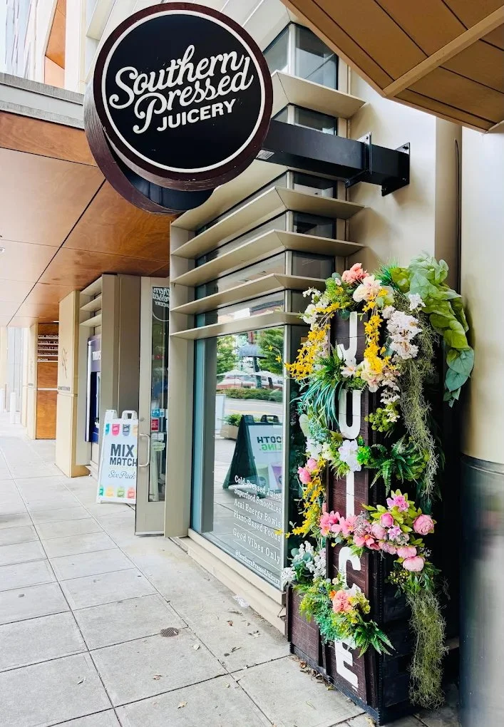 Exterior view of Southern Pressed Juicery with a round black signboard and a colorful floral display on a black vertical sign next to the entrance.