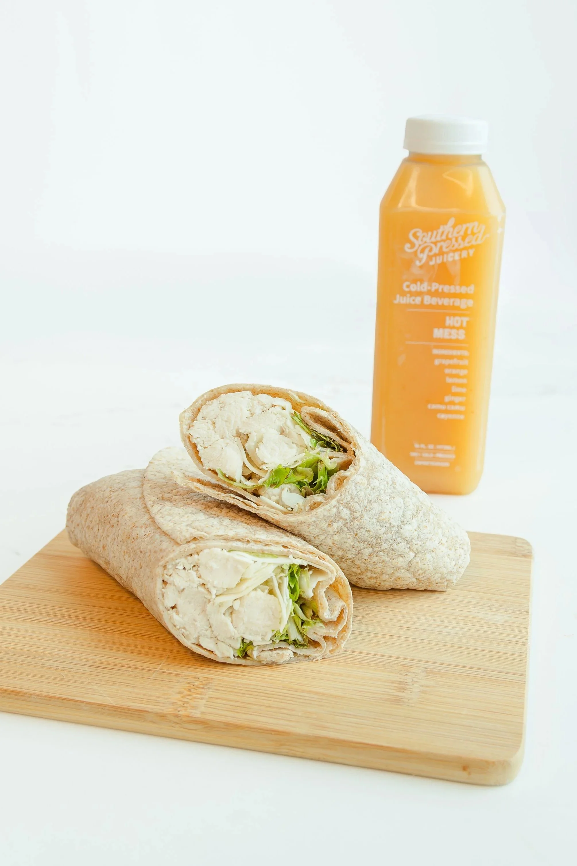 Two wrap sandwiches filled with chicken, lettuce, and cheese on a wooden cutting board, with a bottle of orange juice labeled "SouthernPressed Juicery Cold-Pressed Juice Beverage" in the background.