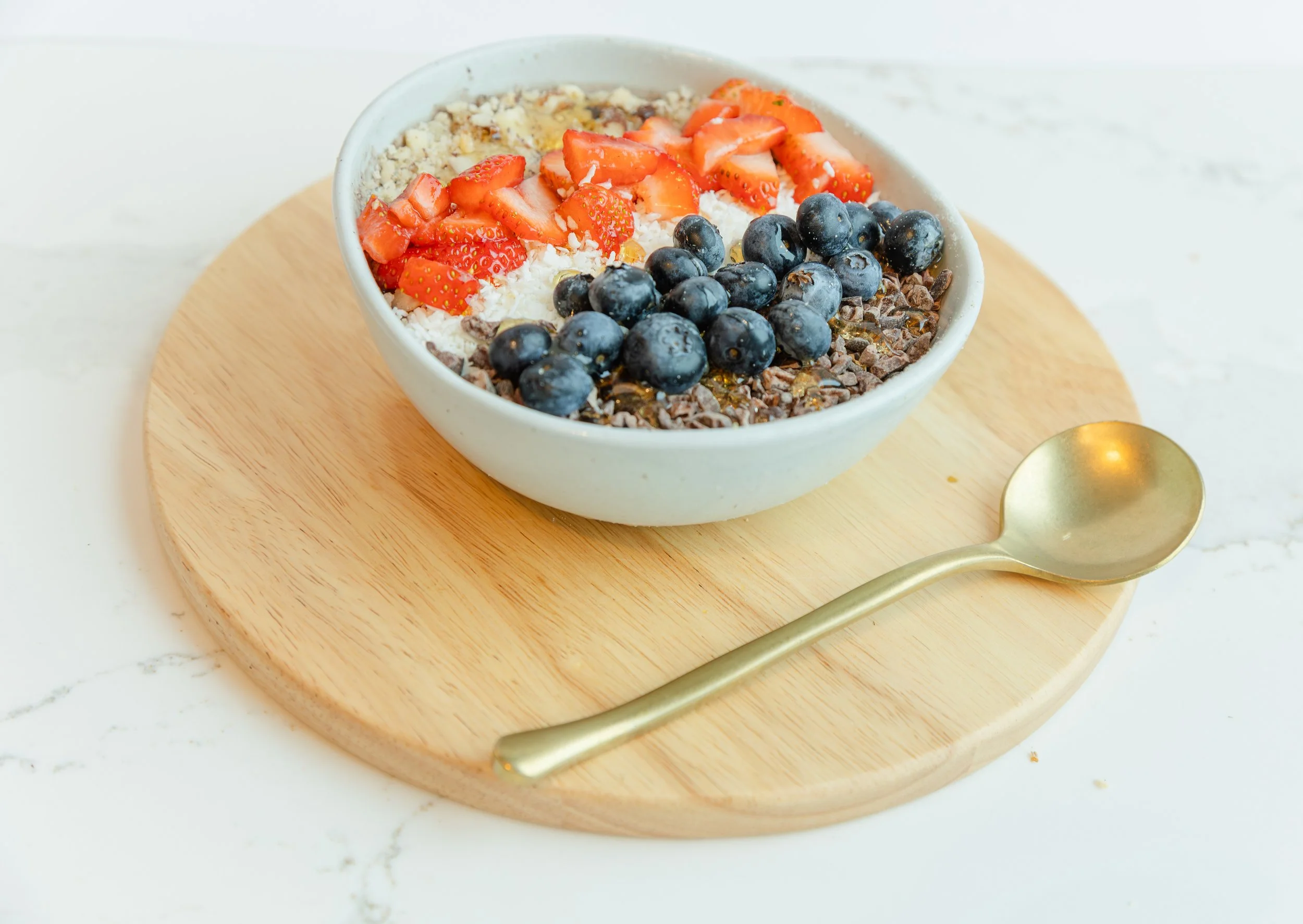 A bowl of yogurt topped with sliced strawberries, blueberries, shredded coconut, and chocolate shavings on a round wooden cutting board with a gold spoon nearby.