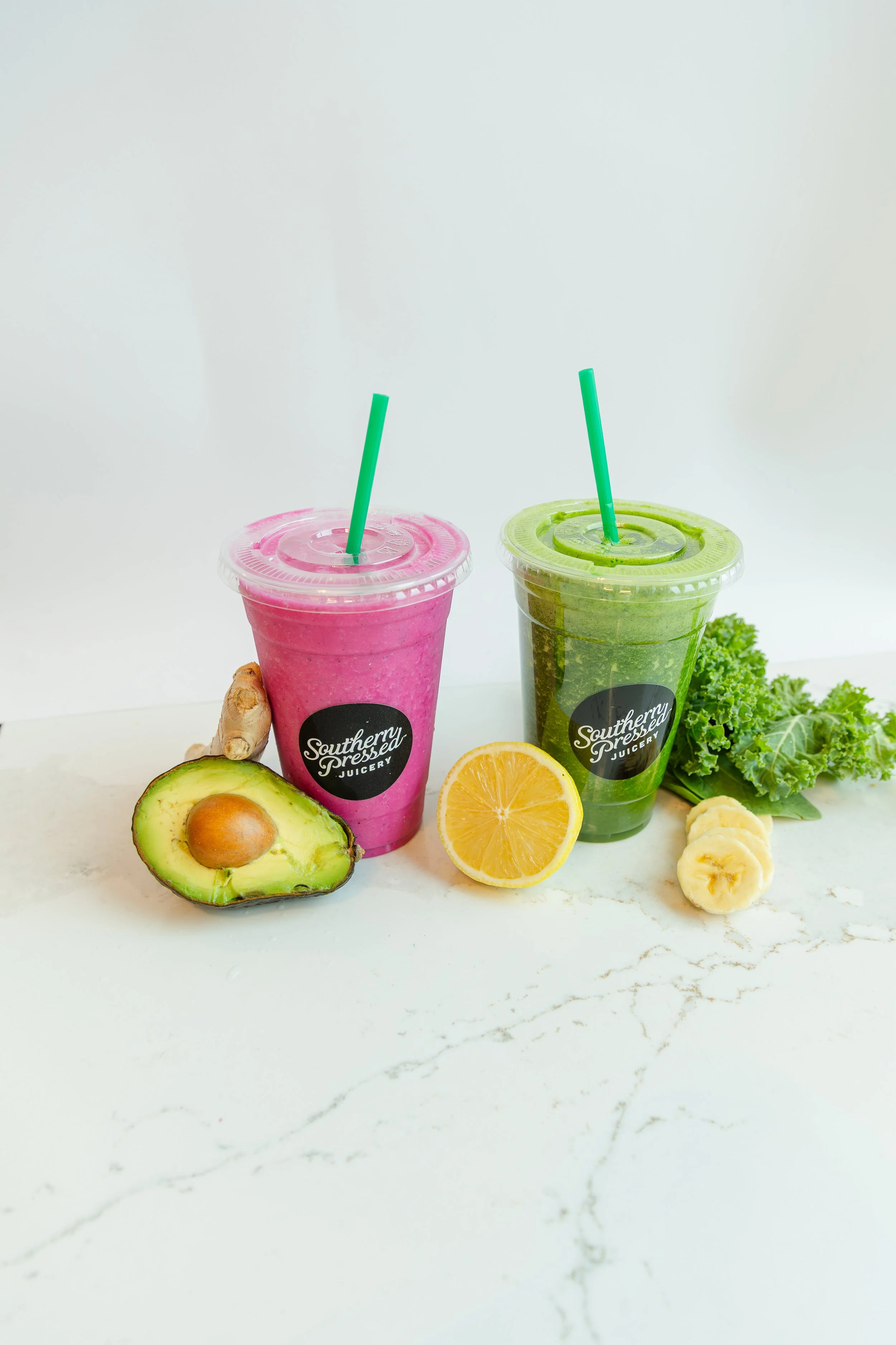 Two smoothies in clear cups with green straws, one pink with avocado and ginger beside it, the other green with lemon, banana, and kale beside it, all on a white surface.