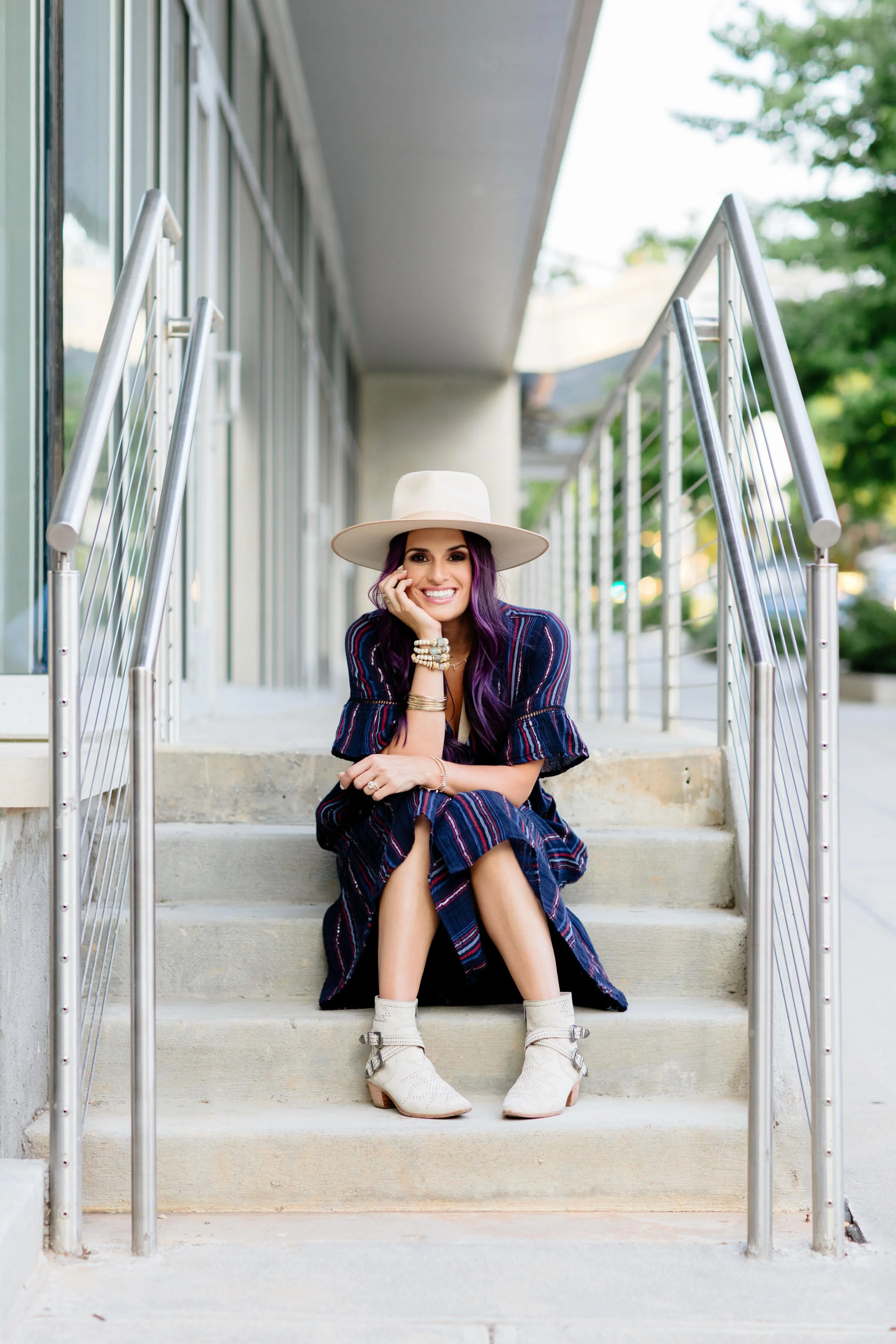 A woman with purple hair, wearing a wide-brimmed white hat, a striped dress, and white ankle boots, sitting on concrete stairs outside a building, smiling at the camera.