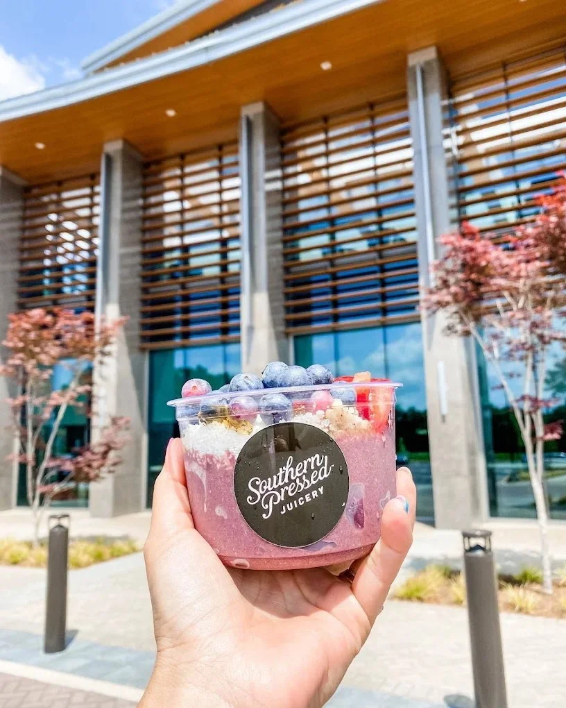 A hand holding a cup of berry smoothie topped with blueberries and strawberries outside a modern building with large windows and wooden slats.
