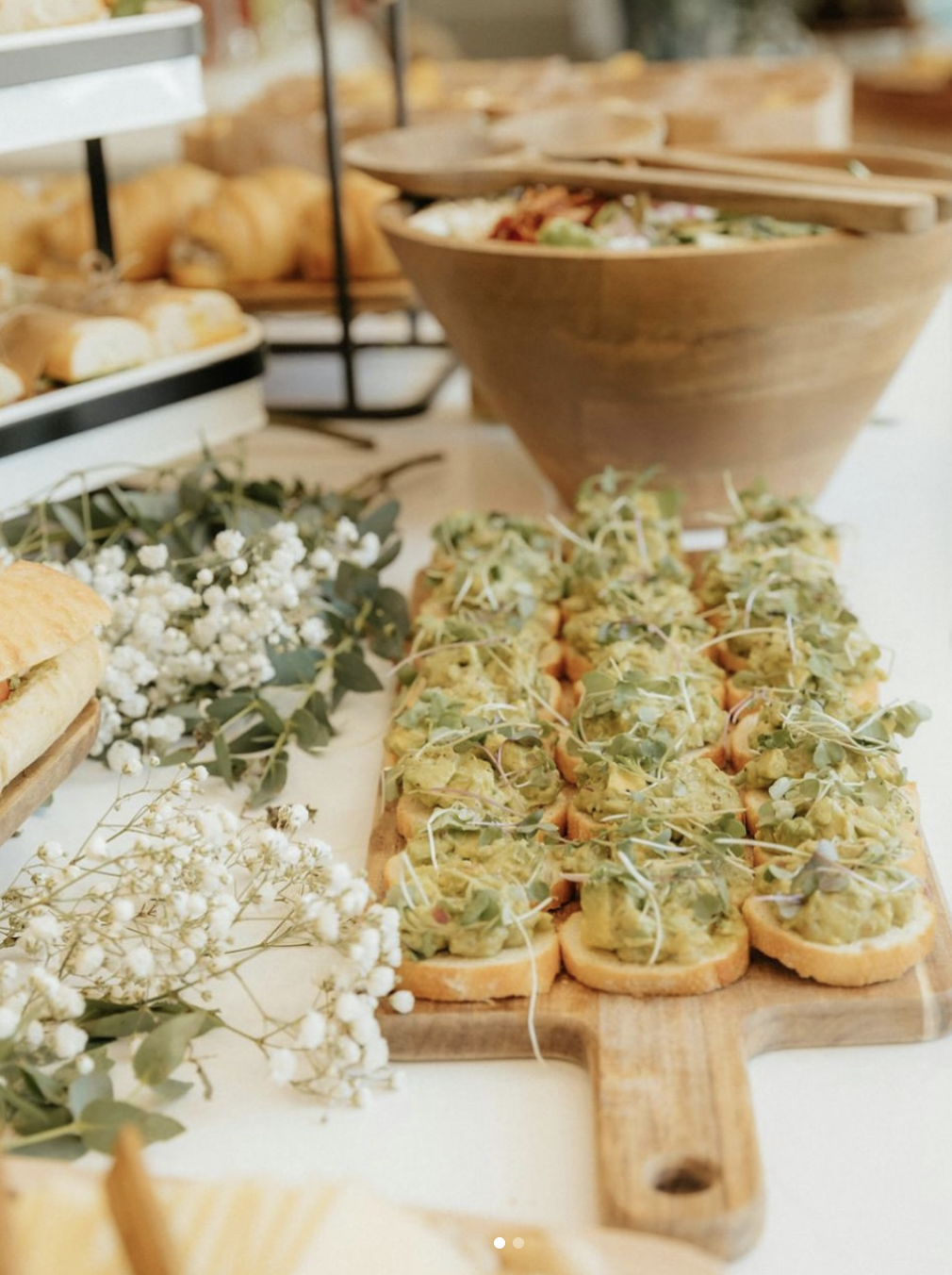 Close-up of a wooden serving board with avocado toast topped with microgreens, surrounded by white flowers, on a white table with other appetizers in the background.