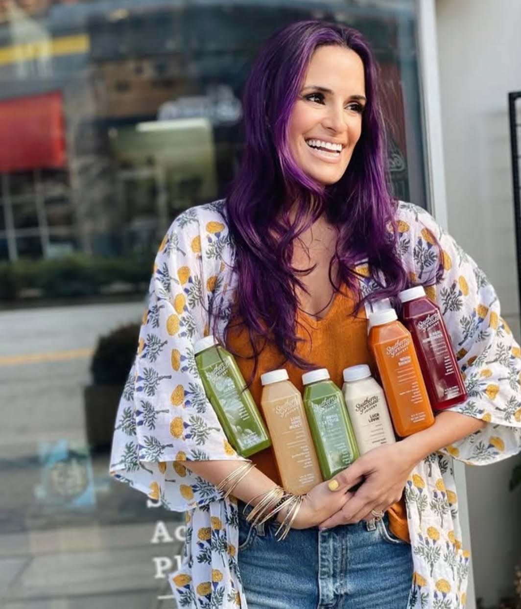 Woman with vibrant purple hair smiling and holding several bottles of juice, dressed in a floral kimono-style jacket over an orange top and blue jeans.