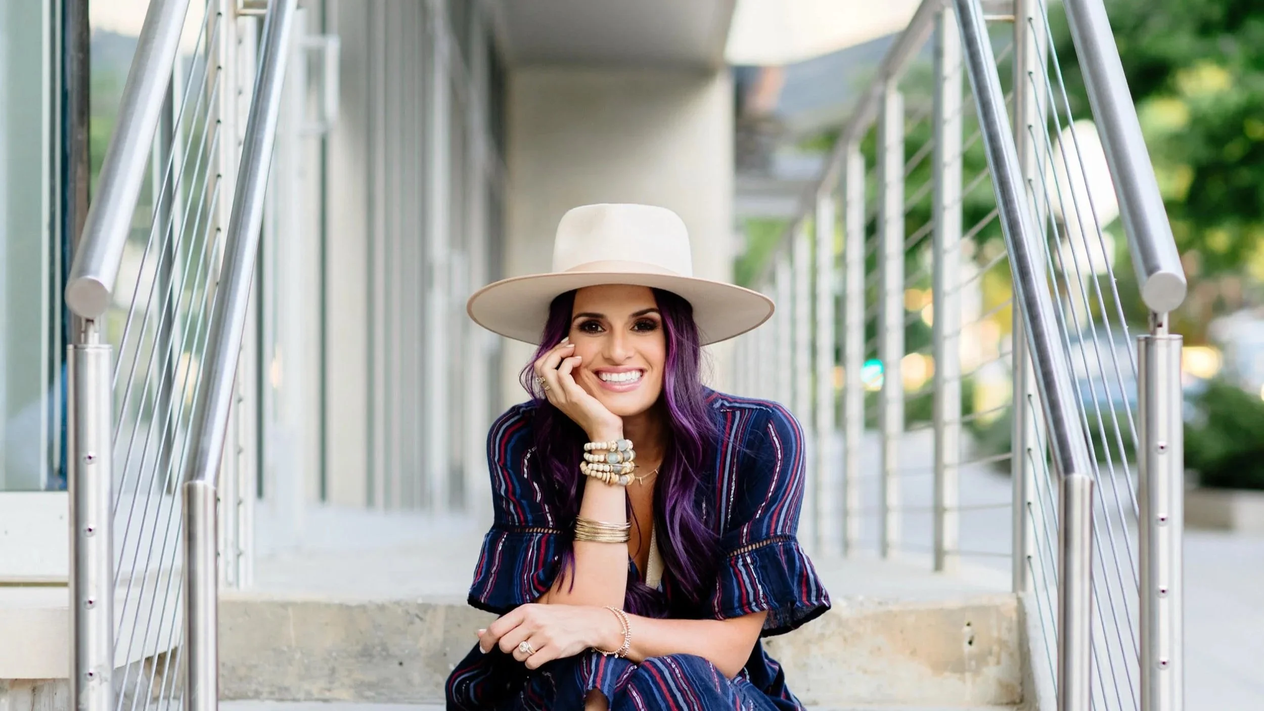 A woman smiling and sitting on outdoor steps, wearing a wide-brimmed hat and striped outfit with purple hair and multiple bracelets.