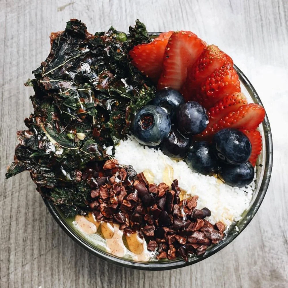 A bowl with kale, strawberries, blueberries, shredded coconut, and chopped nuts on a wooden surface.