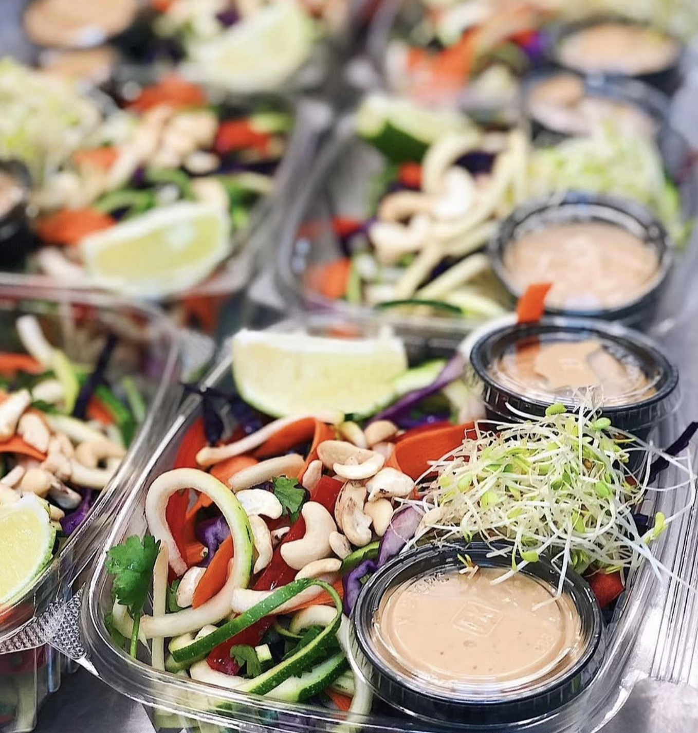 Clear plastic food containers with vegetable salads, lime wedges, and small cups of pink vinaigrette dressing.