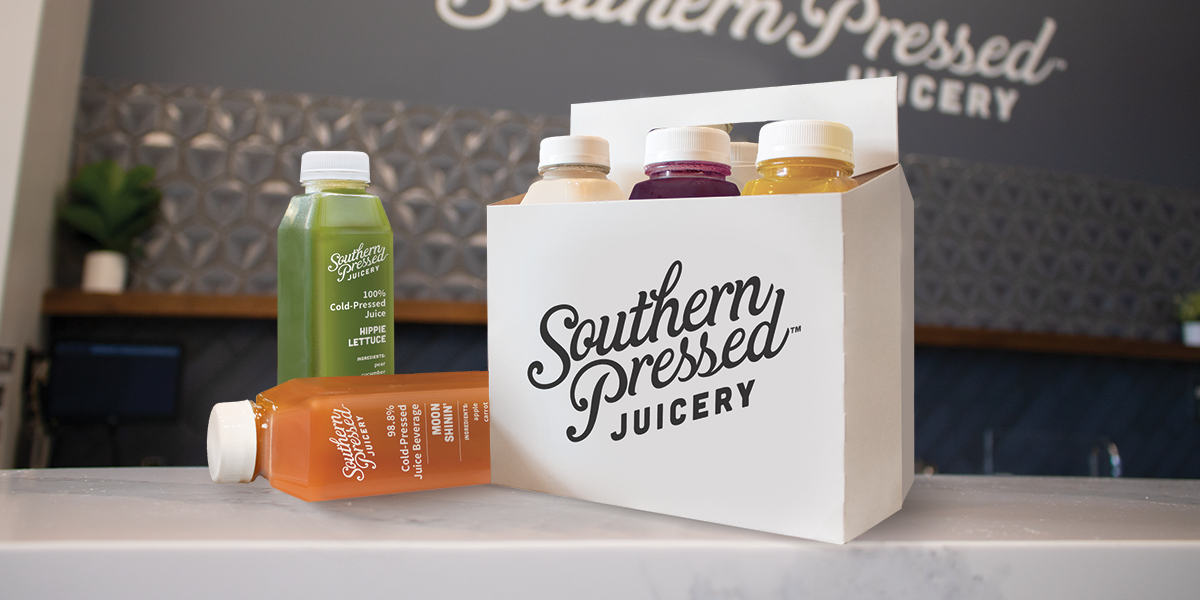 A white paper bag with "Southern Presseed JUICERY" logo, containing four bottles of juice, with an additional bottle lying on the countertop, in a juice shop.