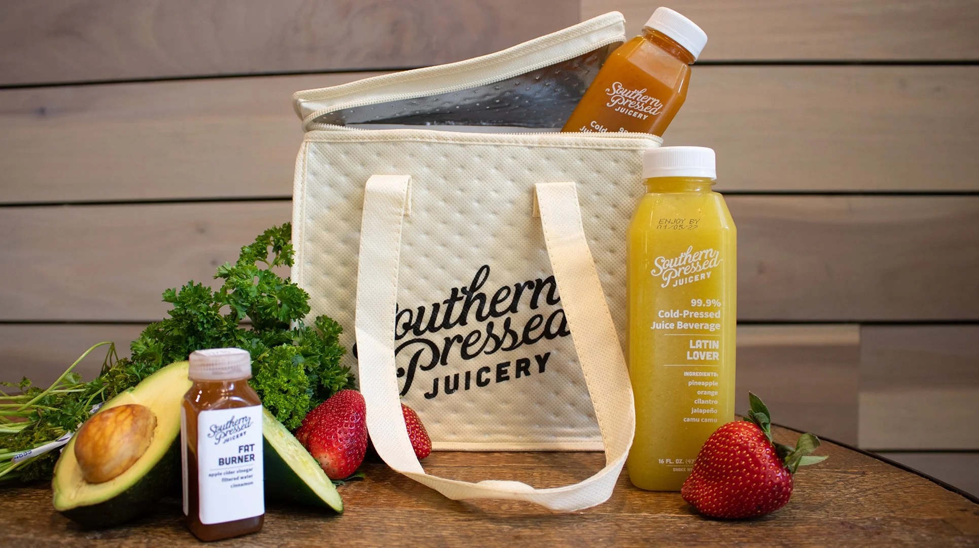 A white insulated bag with black text that reads "Southern Pressed Juicery" is on a wooden surface. In front of the bag are bottles of juice, an avocado cut in half with a seed, strawberries, and fresh herbs. The background features a wooden wall.