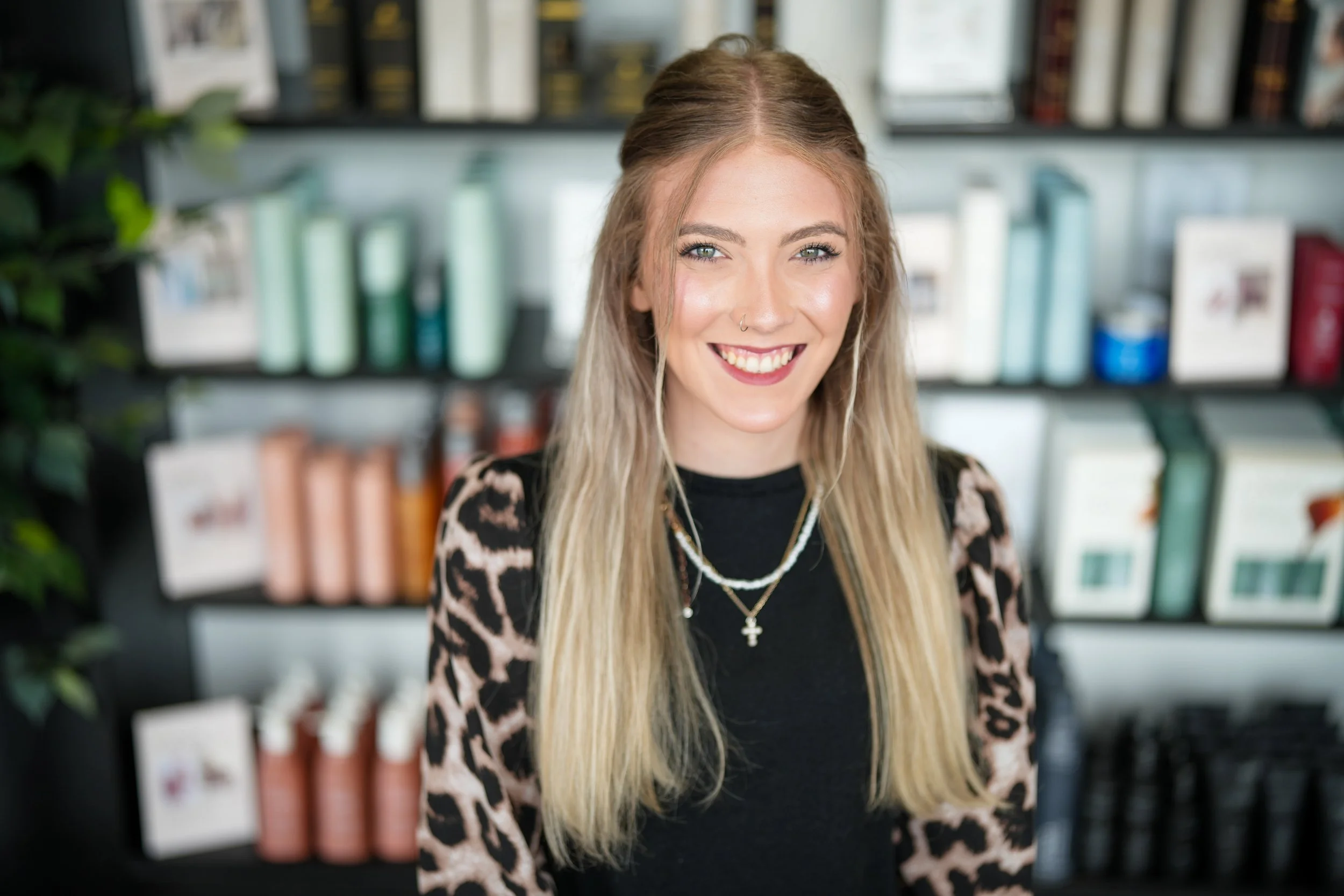 A smiling woman with long blonde hair, wearing a black top with leopard print sleeves and layered necklaces, standing in front of a bookstore or library shelves.