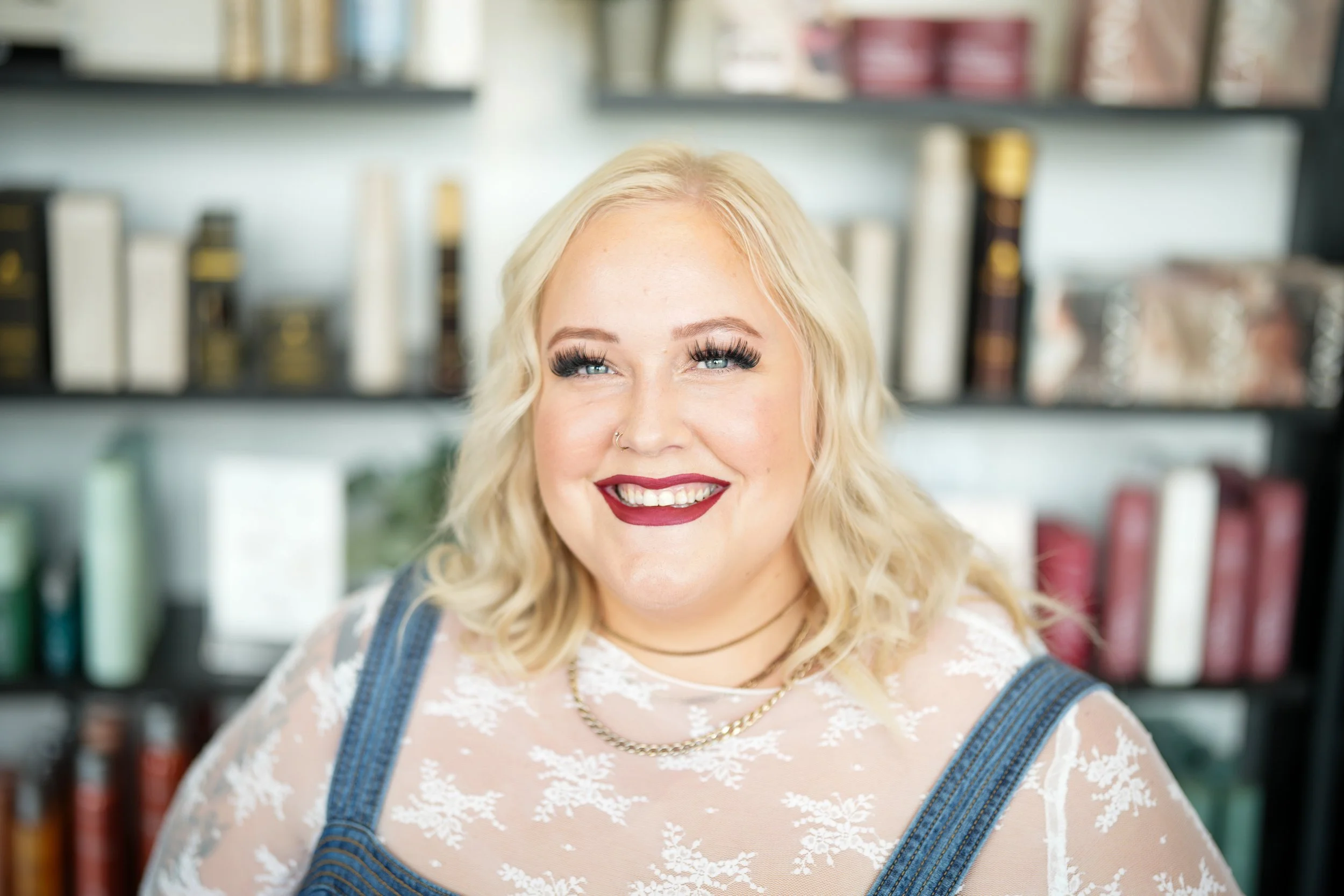 A smiling woman with blonde hair, blue eyes, and makeup, wearing a sheer floral top with denim straps, a gold chain necklace, and a nose ring, standing in front of a bookshelf filled with books.
