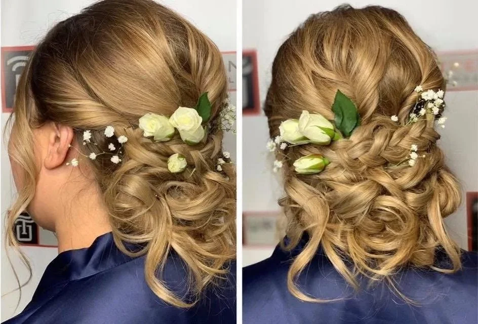 A woman with blonde hair styled in an elaborate updo with loose curls, decorated with white flowers and greenery.
