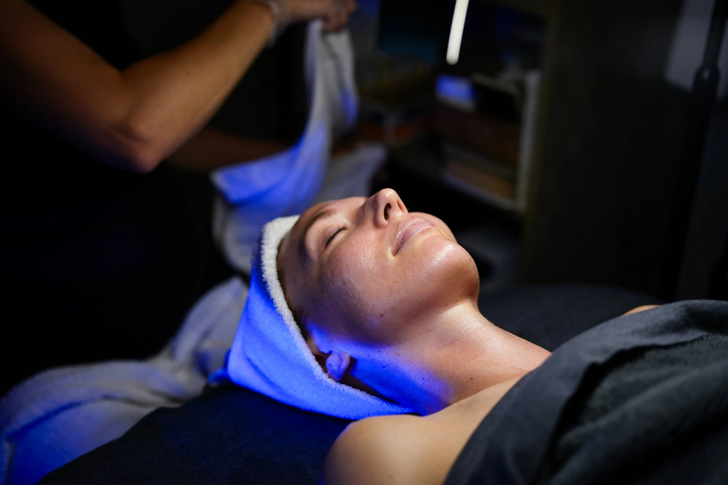 A woman receiving a facial treatment in a spa or skincare clinic, lying down with a towel wrapped around her head, relaxing with her eyes closed.