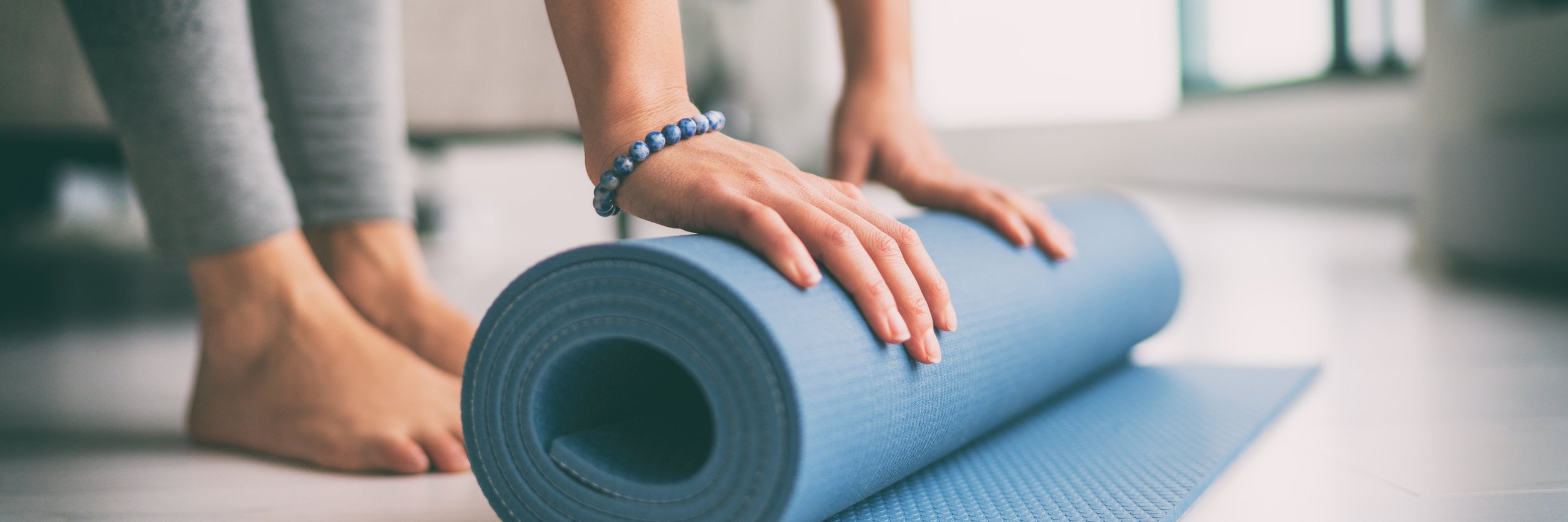 Person unrolling a blue yoga mat on the floor, wearing a bracelet and gray clothing.
