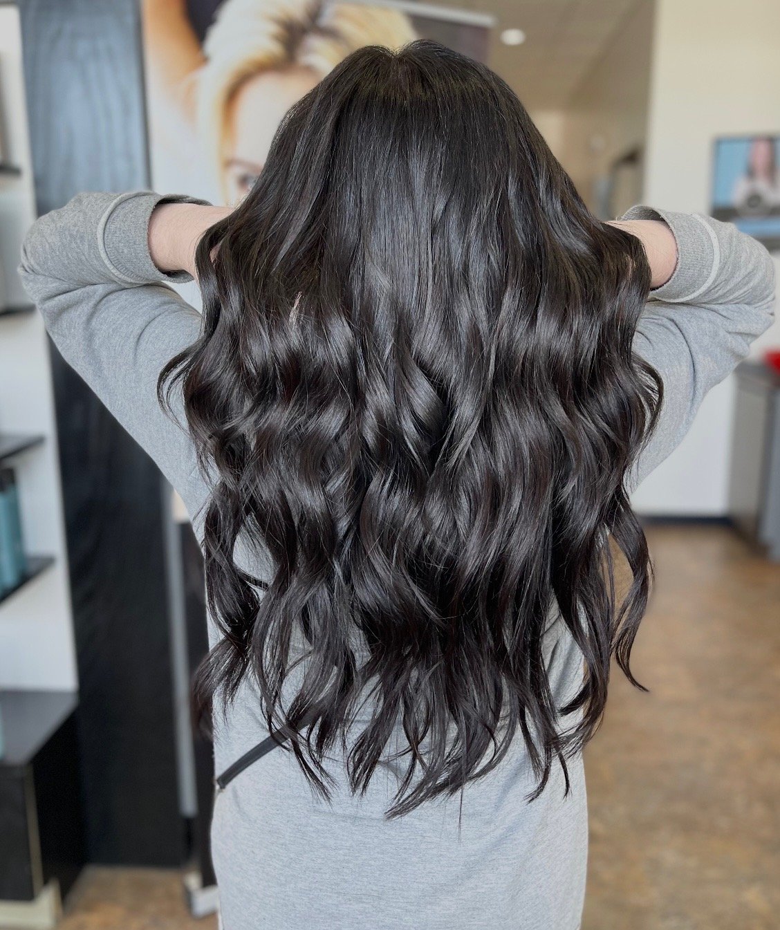 Back view of a woman with long, wavy, dark brown hair, wearing a gray long-sleeve shirt, in a salon or waiting area.
