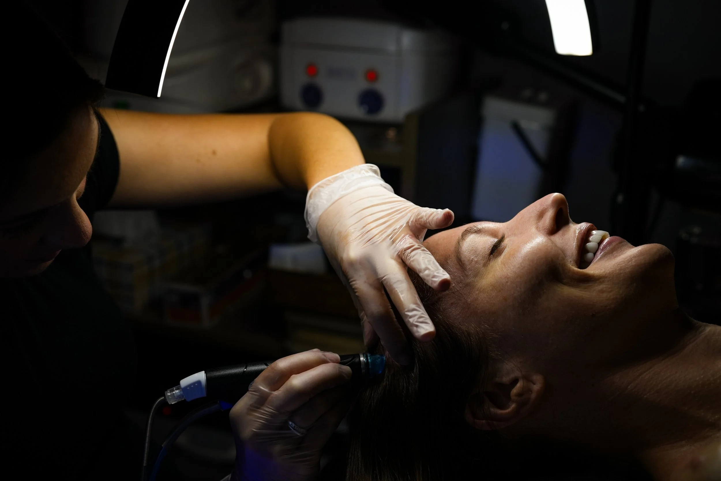 A woman lying back with her eyes closed and smiling while a medical professional uses a device on her head, in a clinical setting.