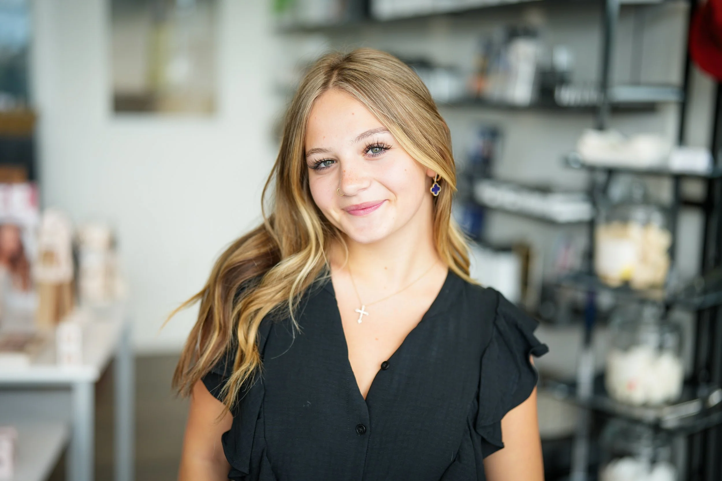 Young woman with blonde hair and a nose ring smiling in a store or cafe with shelves of products in the background.