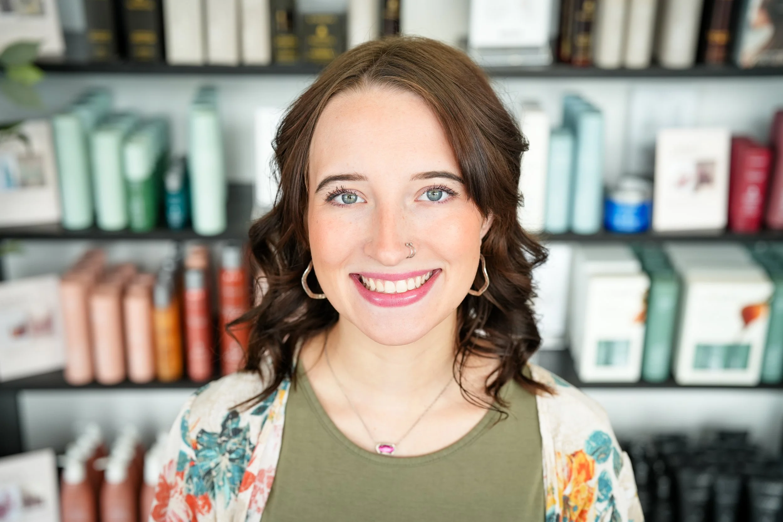 A young woman with brown hair, blue eyes, and a nose piercing, smiling at the camera in front of a shelf filled with various products.