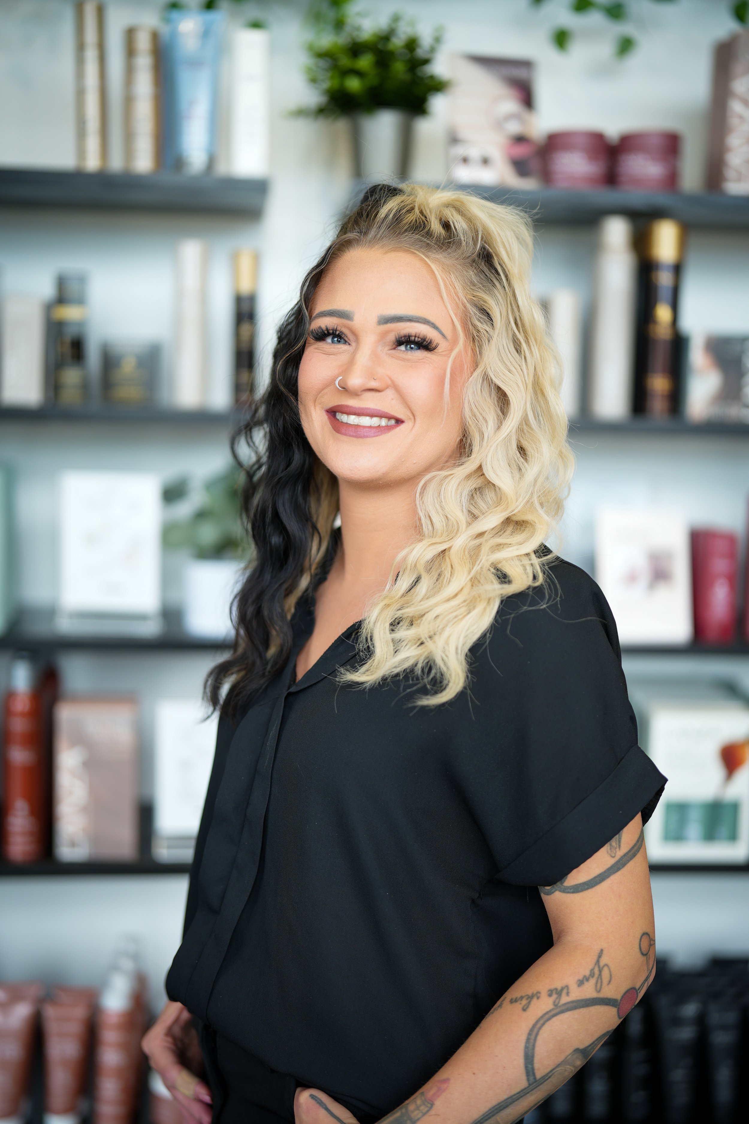 A smiling woman with blonde and black curly hair, wearing a black shirt, standing in front of shelves with beauty products and plants.