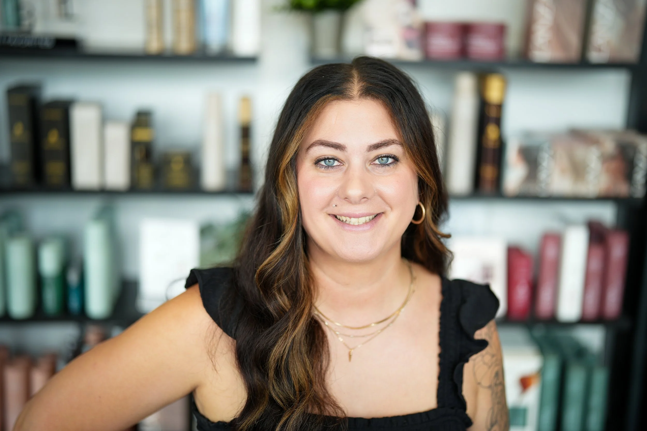 A woman with long dark brown hair, fair skin, and blue eyes smiling at the camera, wearing a black sleeveless top and gold jewelry, with a bookshelf filled with books in the background.