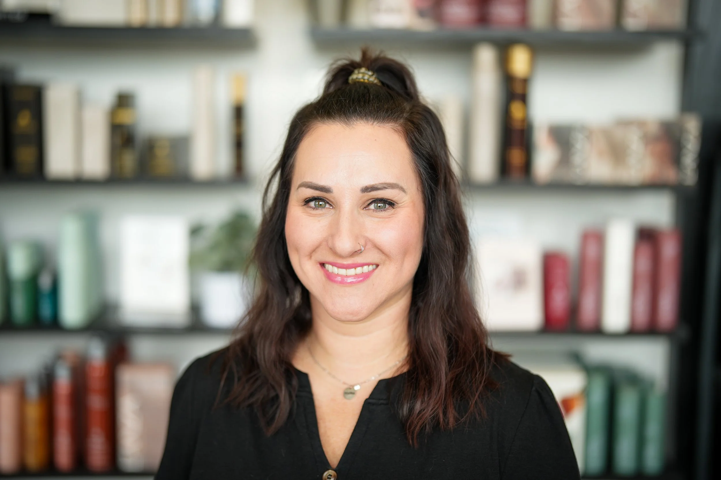 A woman with dark brown hair, light green eyes, and a nose piercing is smiling while standing in front of a bookshelf with books and decor.