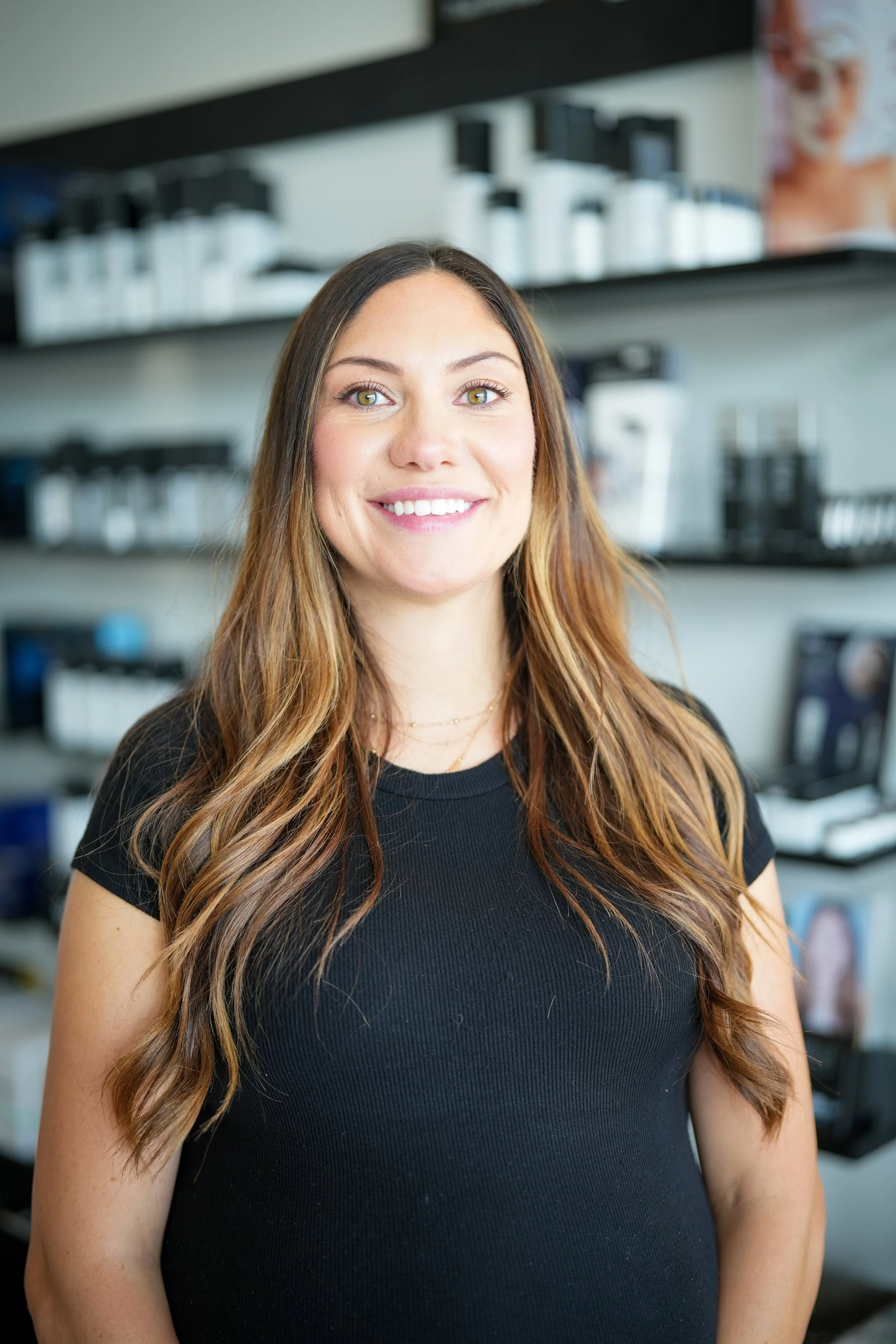 A young woman with long, wavy brown hair smiling, standing in a store with beauty and skincare products on shelves behind her.
