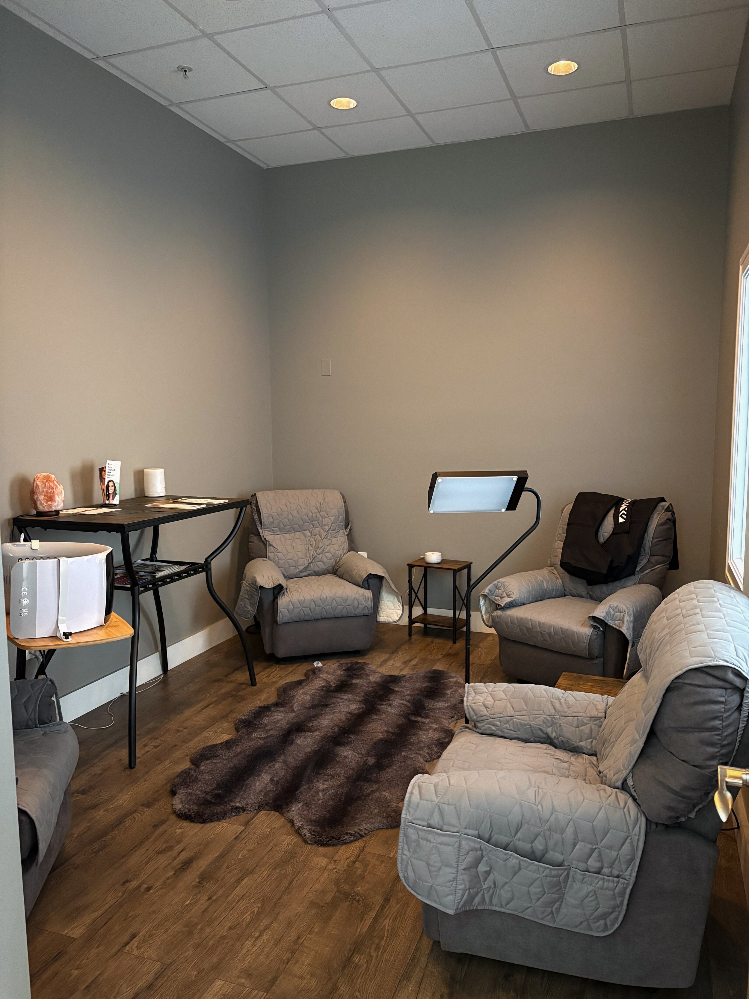 A small waiting room with four grey cushioned armchairs, a dark brown faux fur rug, a small wooden side table, a standing light, and a window. There is a black table with magazines and a salt lamp, and a white diffuser on a small round table.