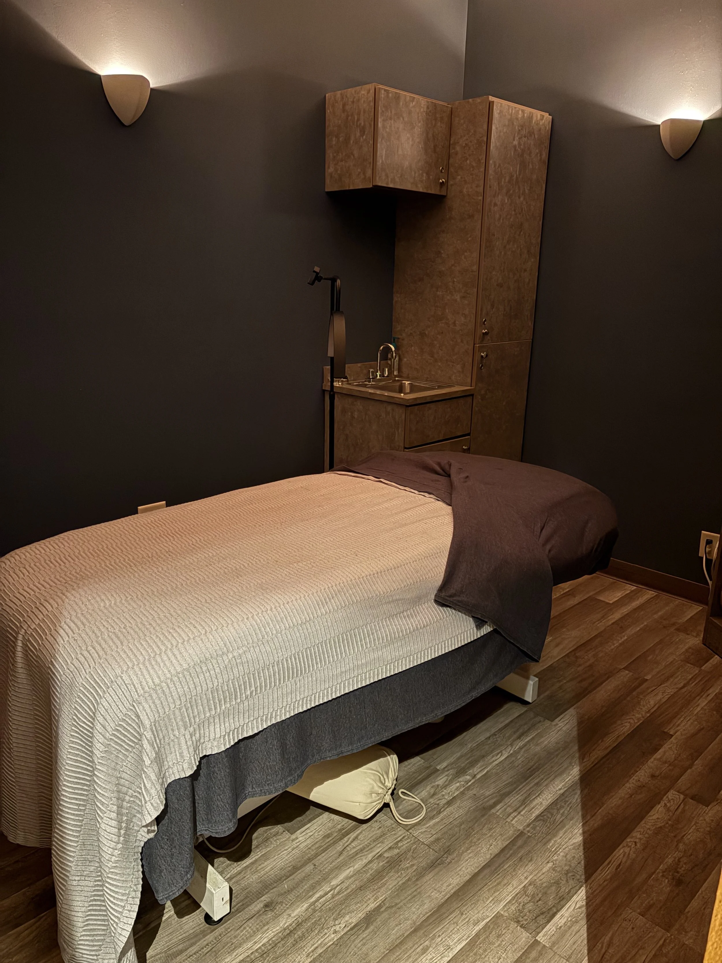 A massage table with a beige sheet and dark brown blanket in a dimly lit room. There is a small sink with a faucet and cabinets in the background, and wall sconces providing soft lighting.
