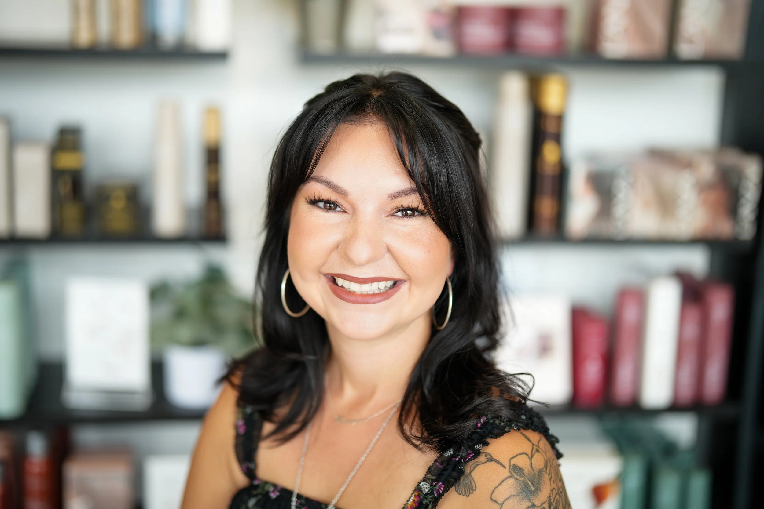 A smiling woman with dark hair, wearing hoop earrings, jewelry, and a black floral top, standing in front of a bookshelf filled with books and decorative items.