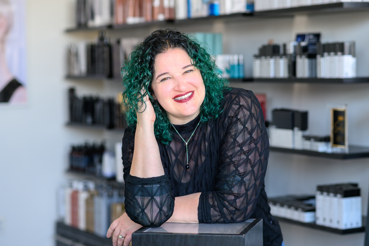 A woman with green curly hair smiling and leaning on a counter in a store or salon, with shelves filled with various bottles and boxes in the background.
