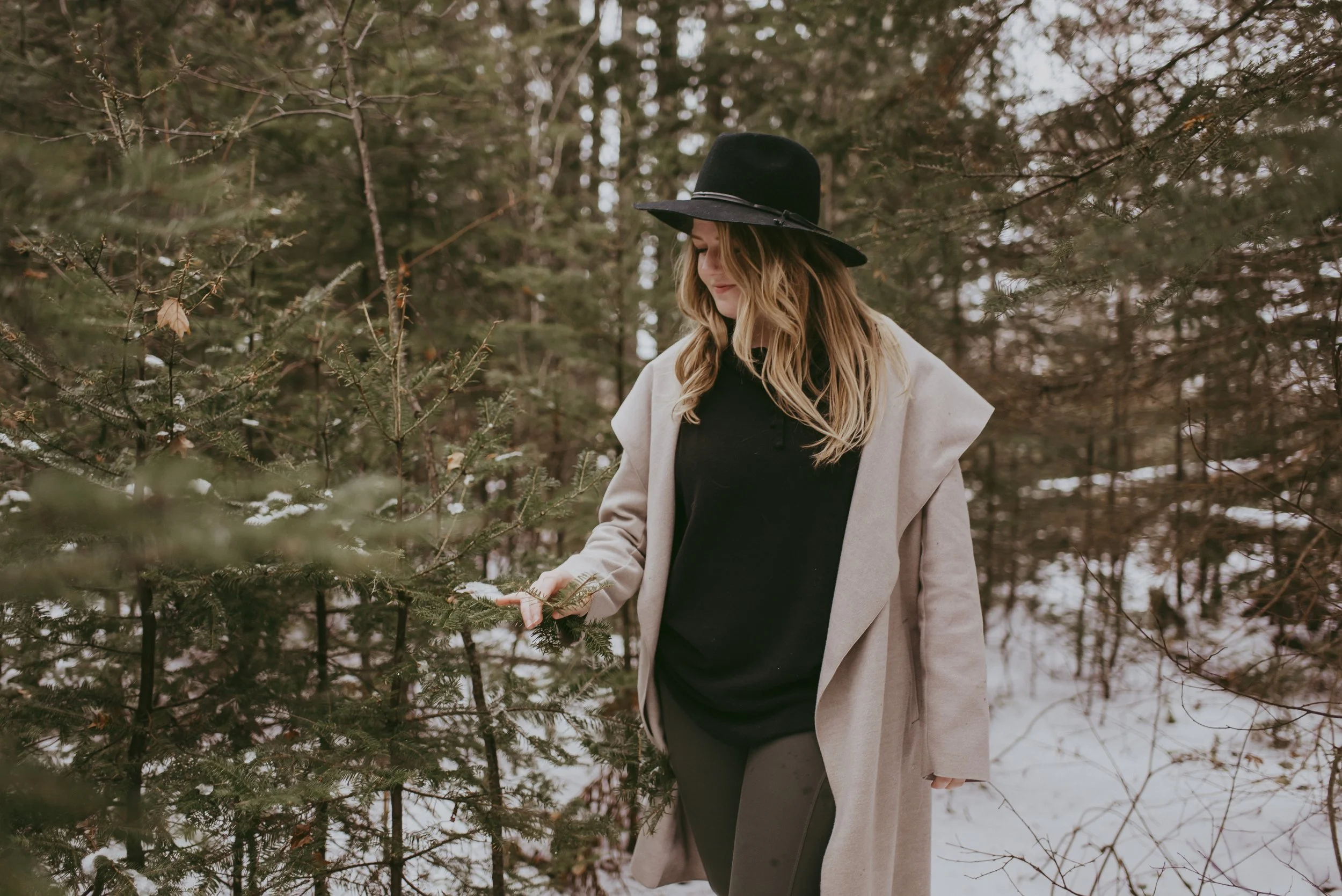 A woman in a beige coat, black hat, and black outfit walks through a snow-covered forest during winter.