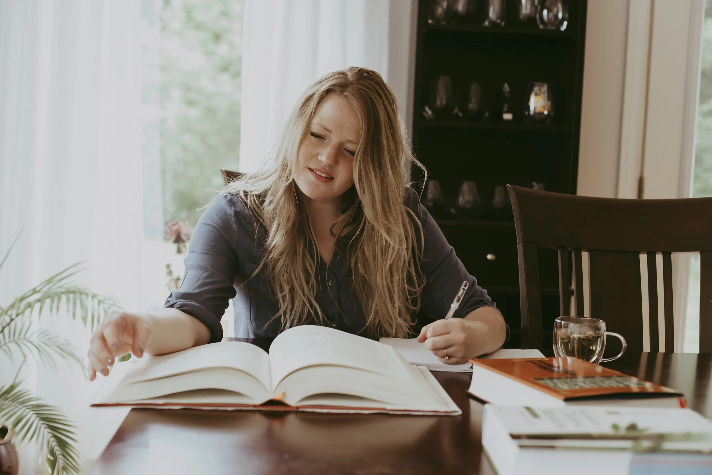 A young woman with long blonde hair, wearing a dark long-sleeve shirt, sitting at a wooden table with open books, a glass of water, and a pen in hand. She appears to be studying or working, with a background of a room with large windows and natural light.