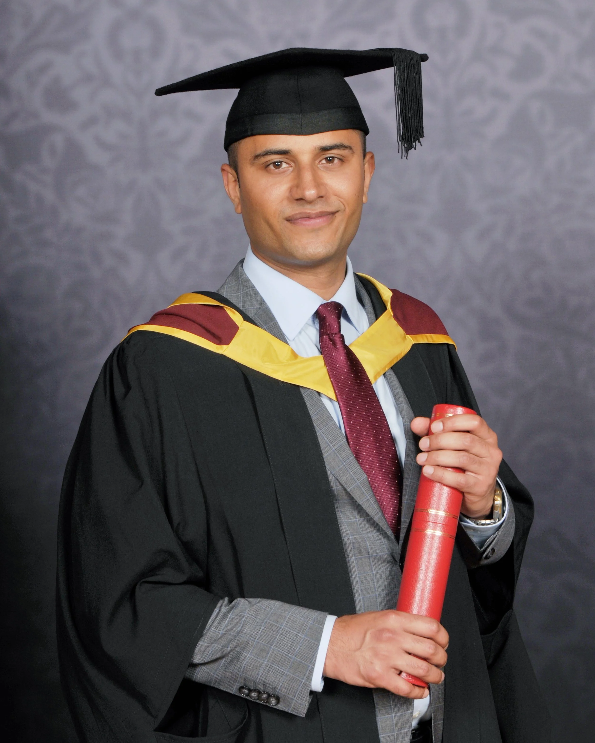 A man in graduation attire holding a diploma, wearing a mortarboard and gown with a hood, smiling at the camera.