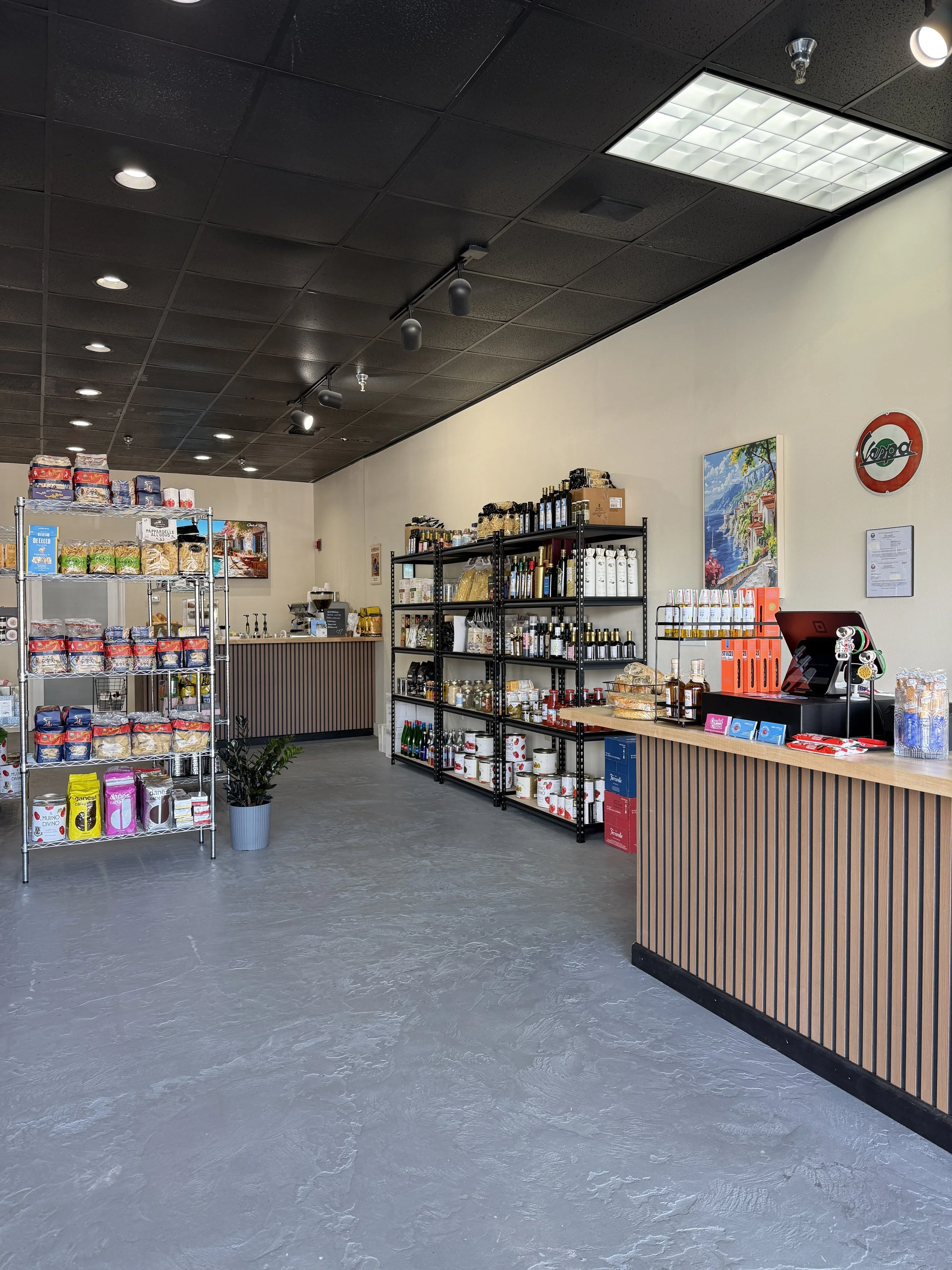 Interior of a small retail store with shelves stocked with various products, a counter in the background, a wall with a colorful landscape painting, and a gray floor.