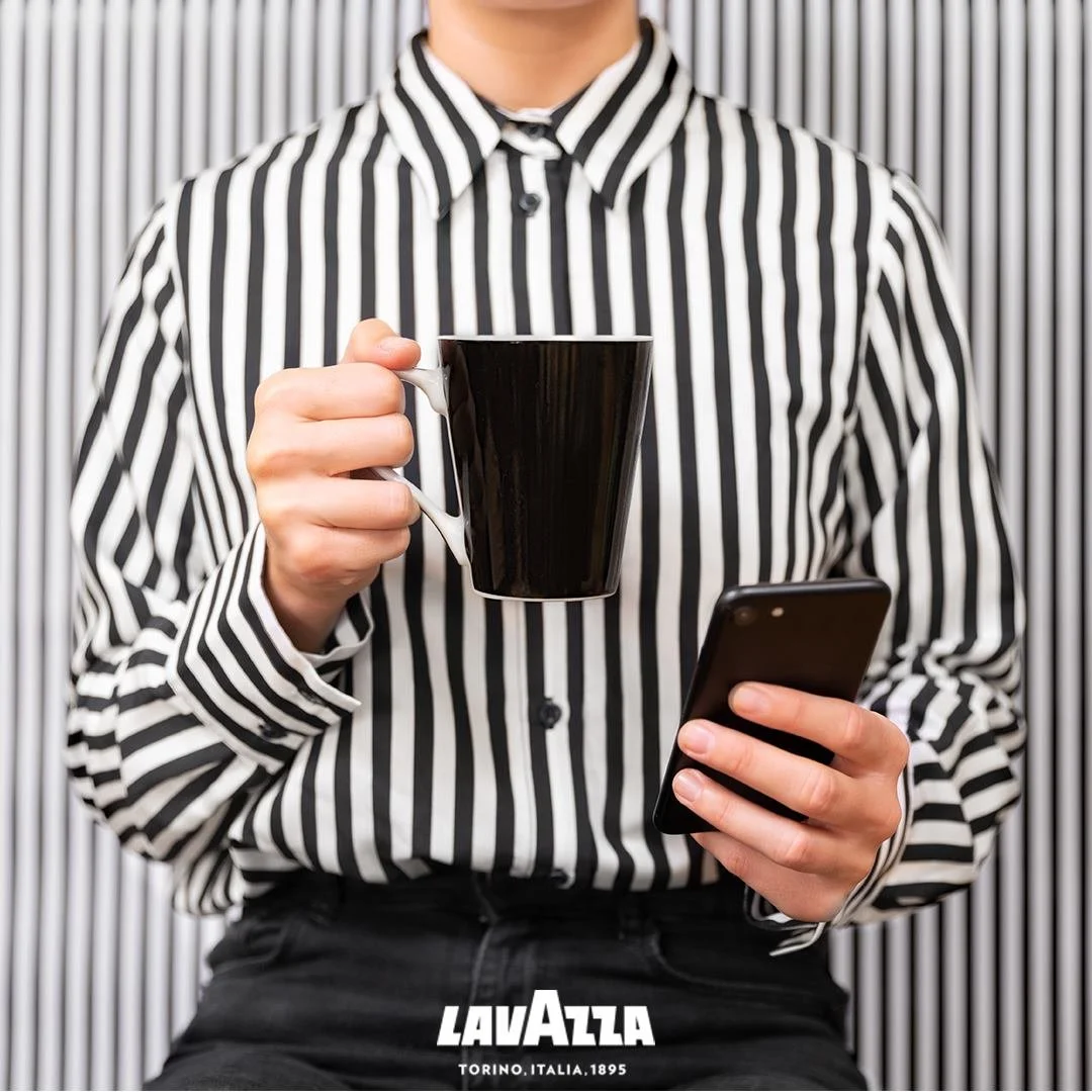 Person wearing a black and white striped shirt holding a black coffee mug in one hand and a smartphone in the other, with vertical striped background and Lavazza logo at the bottom.