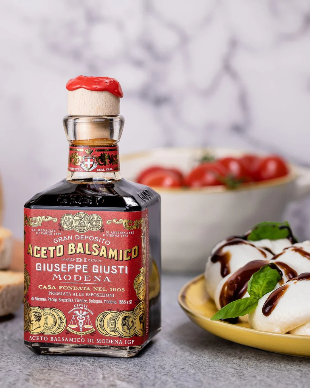 Bottle of balsamic vinegar with a red label, next to a dish of mozzarella cheese drizzled with balsamic glaze and fresh basil, and a bowl of cherry tomatoes in the background.