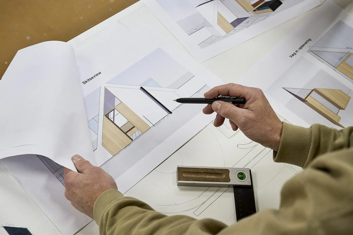Person der arbejder med arkitektoniske tegninger af et hus, med farvede materialer og skitseoversigter.