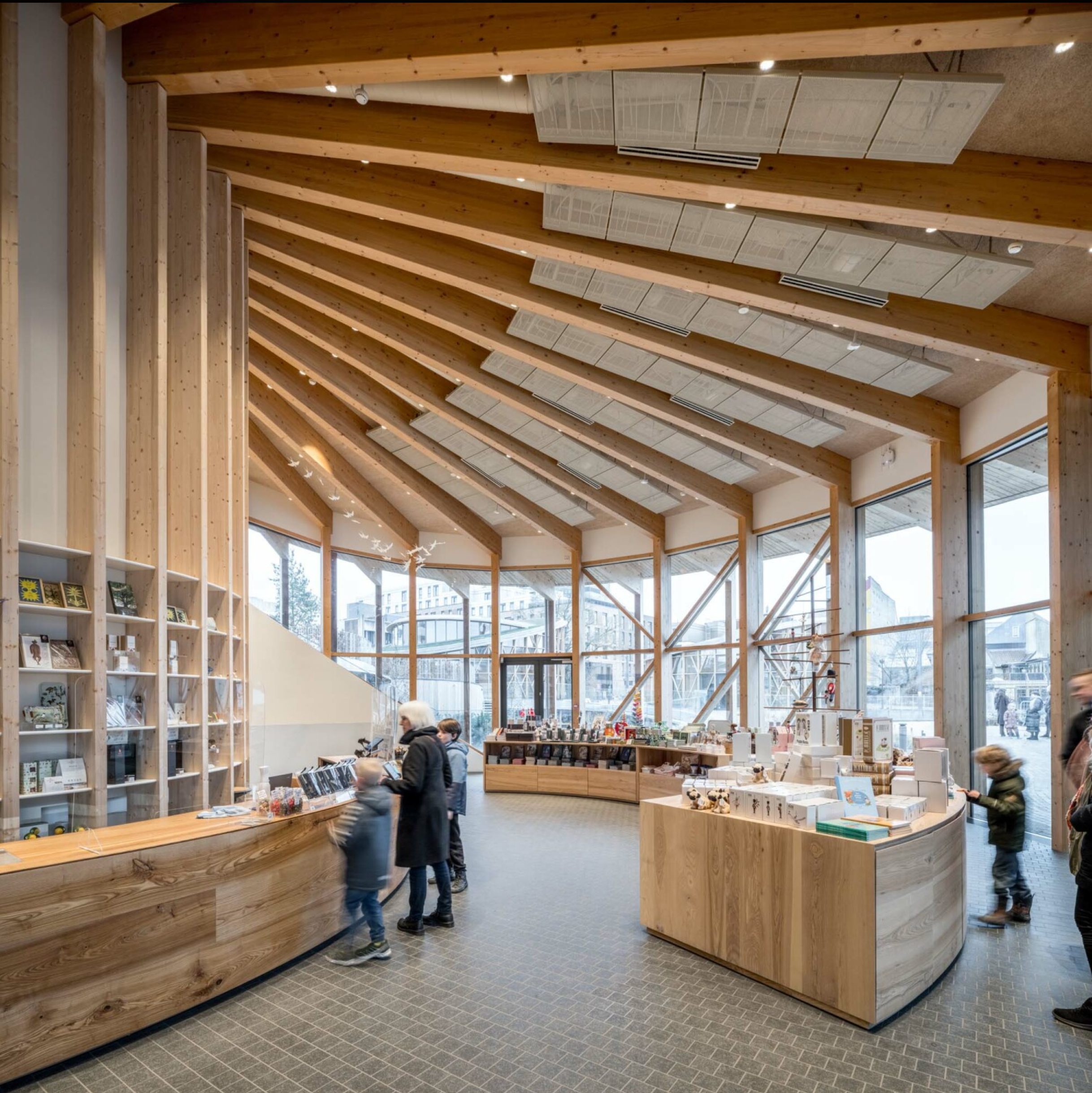 Indendørs område med træmaterialer og borde, børneshoppe med legetøj og bøger, store vinduer, mange mennesker.