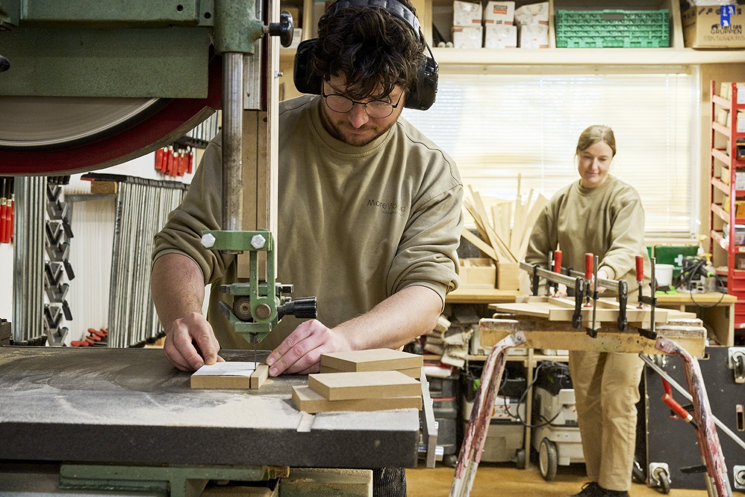 To personer arbejder i en træværksted. Den person i forgrunden bruger en bordsav til at skære i træ, mens den anden person i baggrunden står ved et arbejdsbord med træstykker og bruger klammer til at fastgøre materialer.
