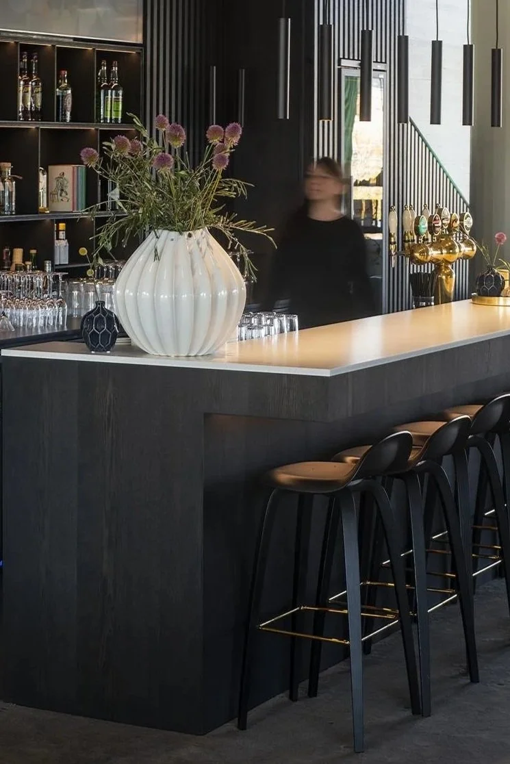 Indendørs barområde med sort træudskæring, barpult med hvide bordplader, store blomster i en hvid vase, samt alkoholholdige drikkevarer på hylderne. En bartender bevæger sig bag baren, og der hænger moderne sorte lamper ned over baren.