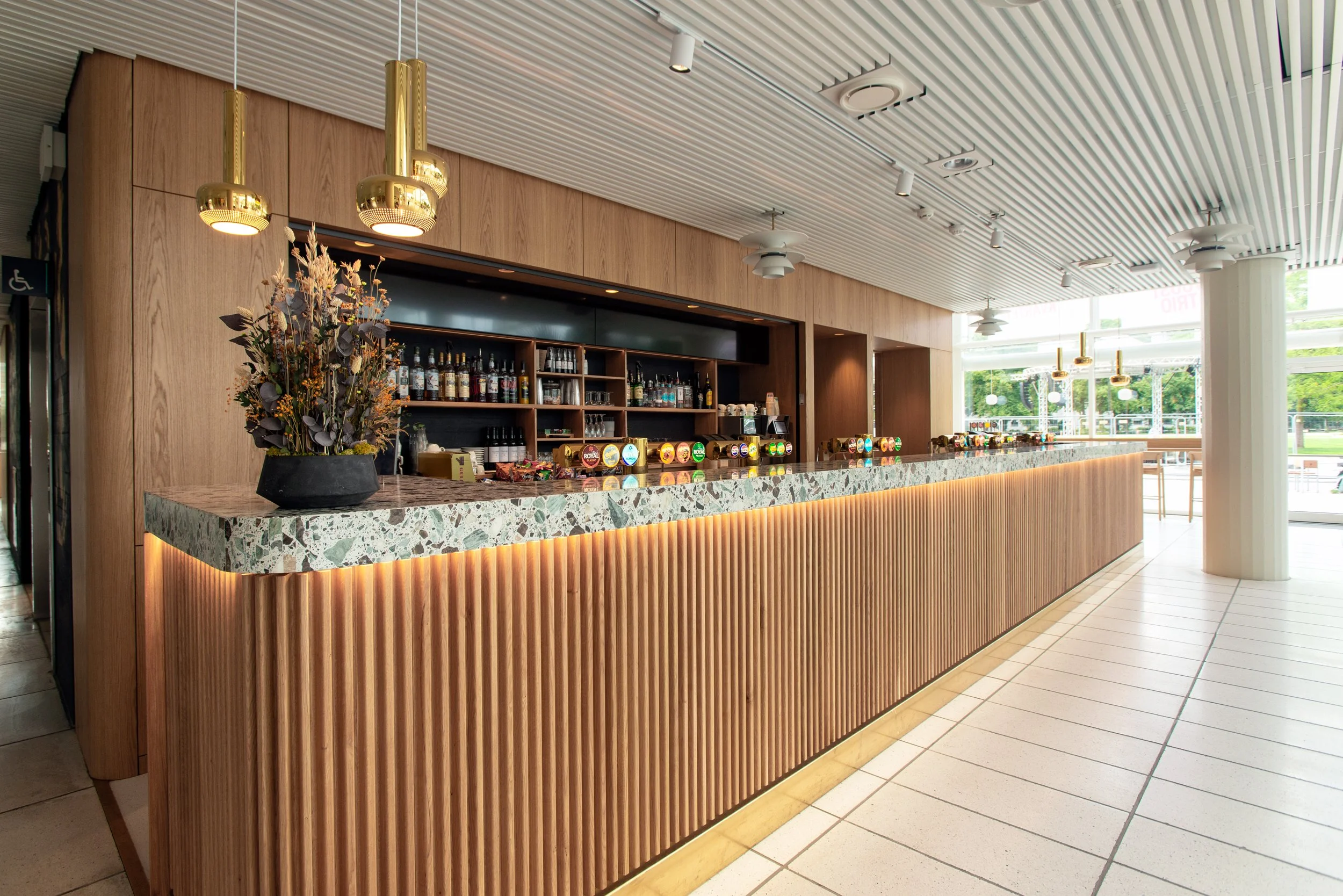 Indendørs billede af en bar med træpanelvægge og en terrazzo-borddisk, hvortil der er anbragt en blomsterdekoration. Der er store vinduer bag baren, der slipper naturligt lys ind, og lyse loftinstallationer.