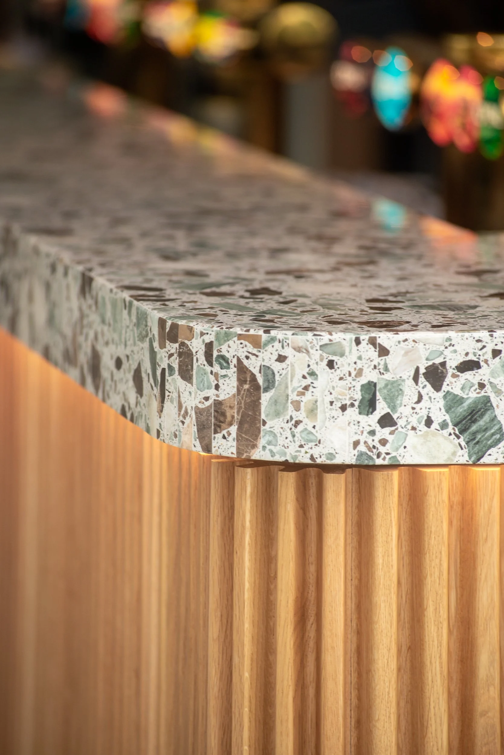 Et køkkenbord lavet af terrazzo med en trælampe under bordet, og en baggrund med farverige lysreflektioner og udskårne vægge.