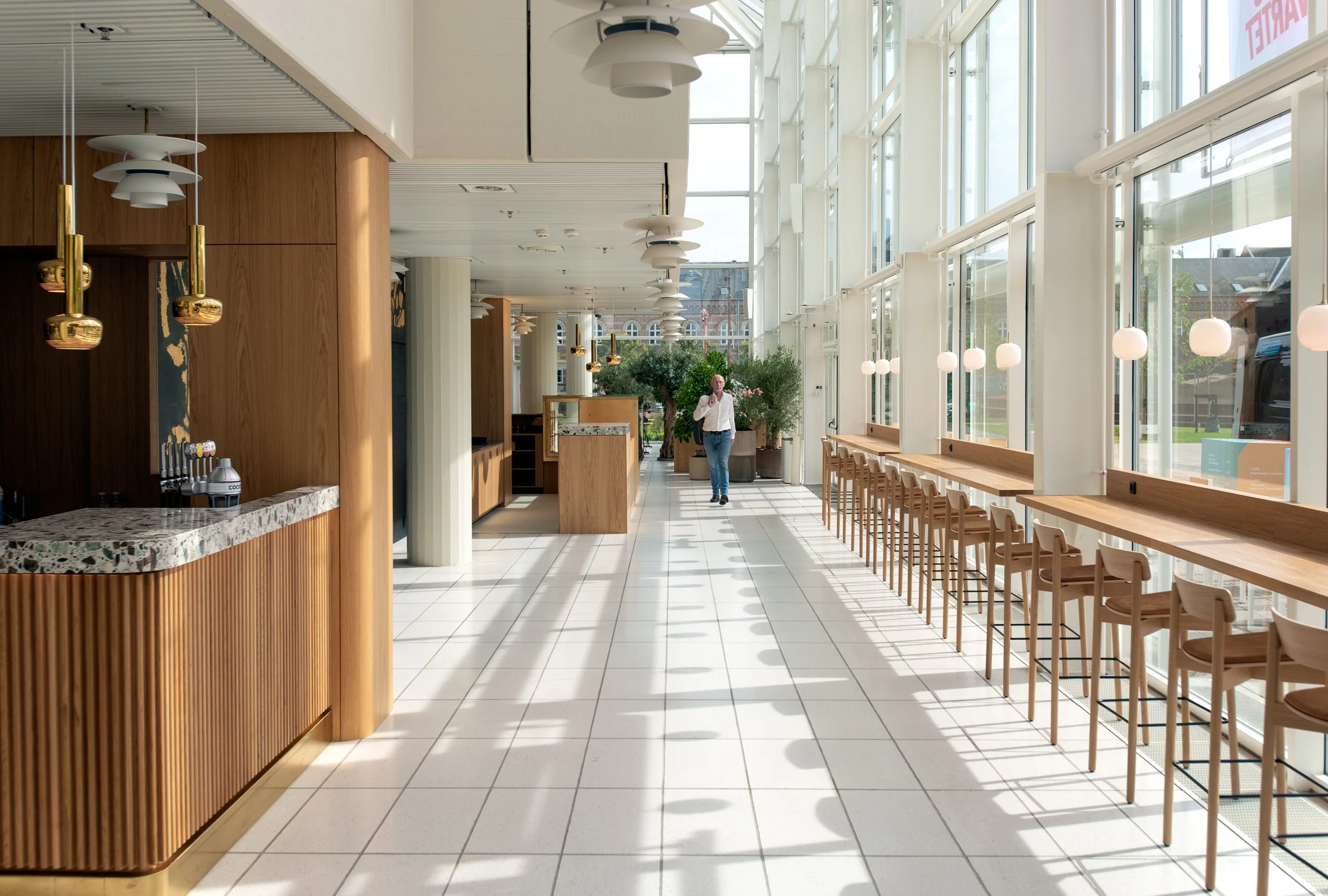 Indendørs café med store vinduer, træborde med stole, hængende lamper og planter, en person går i baggrunden.
