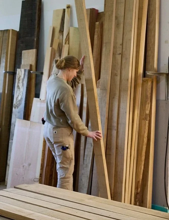 En kvinde i beige tøj står i en træbutik eller værksted, der er omgivet af forskellige stykker træ og planker, mens hun rører ved en stor lang træbjælke.