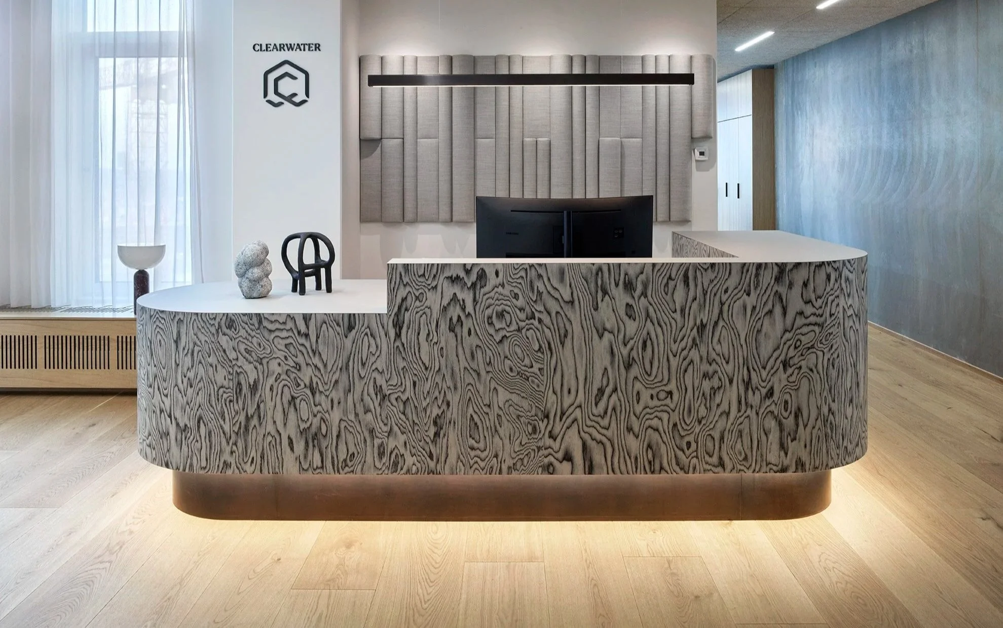 Moderne reception med en krøllet træskranke, dekorative genstande, sorte stole og en bagvæg med lyse vægpaneler i en lys, rummelig indretning.