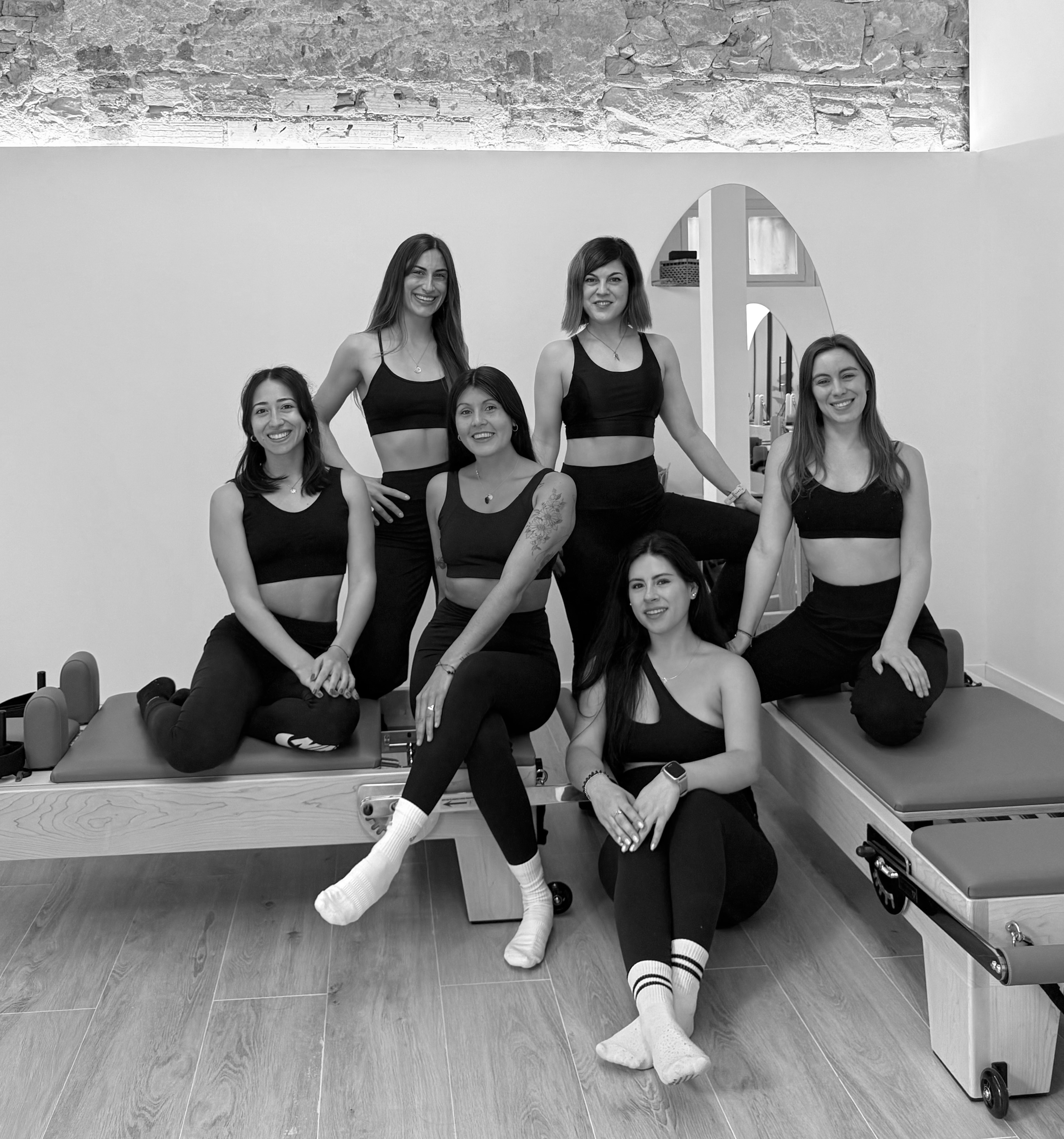 Grupo de siete mujeres en ropa deportiva posando en un estudio de fisioterapia o rehabilitación, algunas sentadas y otras de pie, en un ambiente con paredes blancas y fondo de muro de ladrillos.