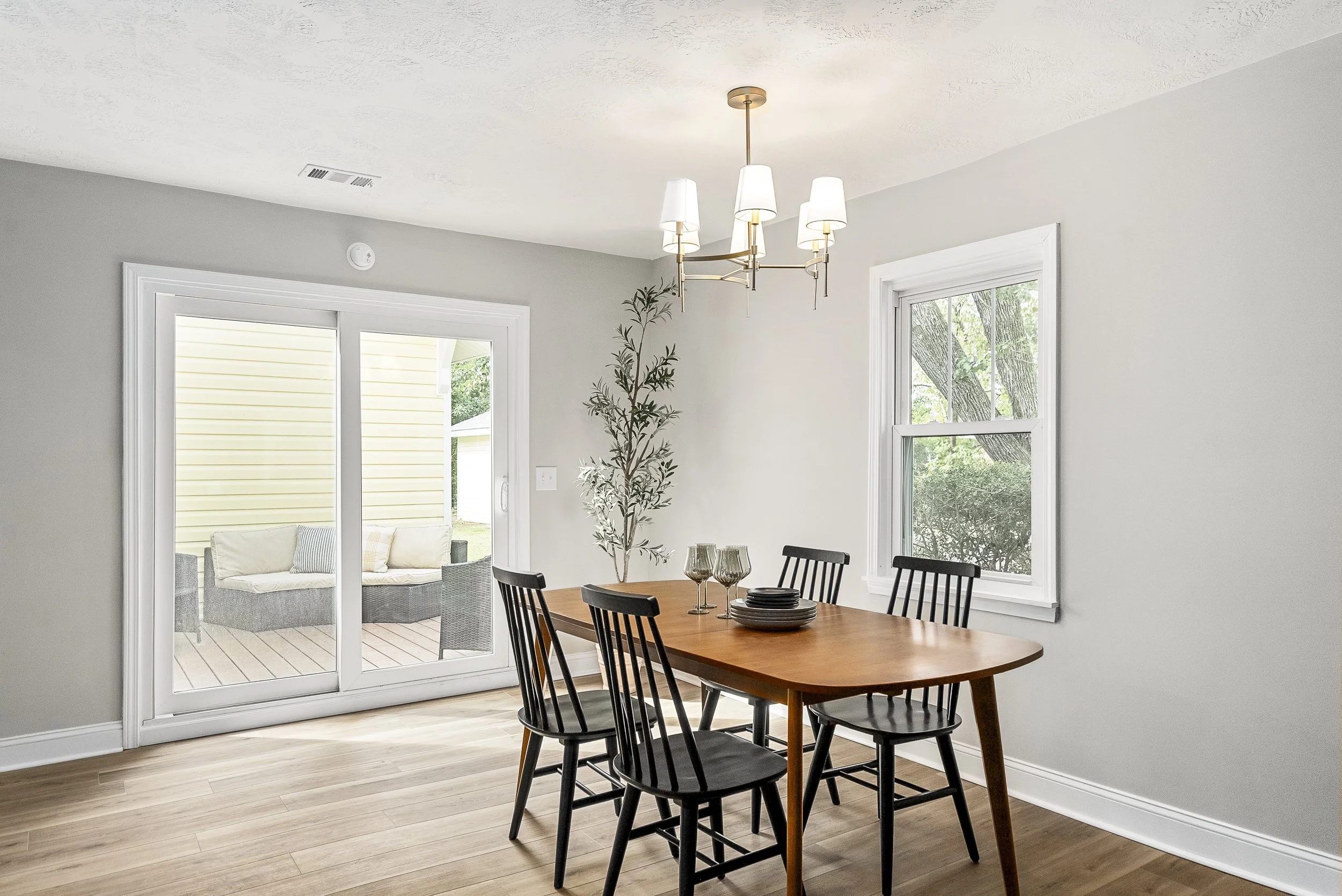 A dining room with a wooden table, black chairs, and a chandelier hanging from the ceiling. There are large windows letting in natural light, and an outdoor patio with a seating area is visible through sliding glass doors.