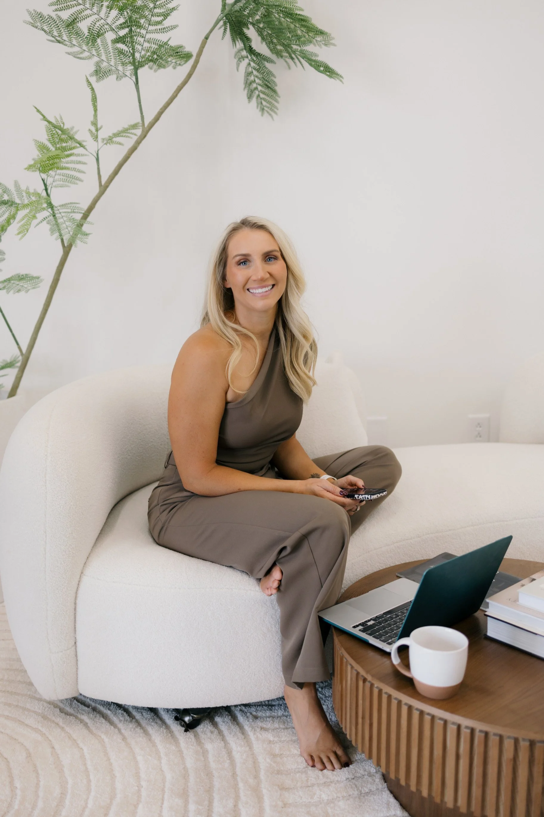 A woman with blonde hair, wearing a taupe sleeveless top and taupe pants, sitting barefoot on a white curved sofa, smiling and holding a remote control, in a modern living room with a coffee table, an open laptop, a coffee mug, a stack of books, and a large green leafy plant in the background.